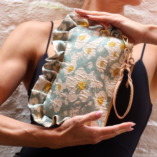 The Dolly Blurred Daisies Frilly Edge Bag is handcrafted in England from elegant mint green, cream and yellow jacquard, finished with a caramel moiré lining, yellow gold zip and gold hardware. Complete with a natural leather wrist strap and bespoke gold chain, it can be worn as a clutch, shoulder or crossbody bag for weddings, festivals, holidays and everyday occasions.