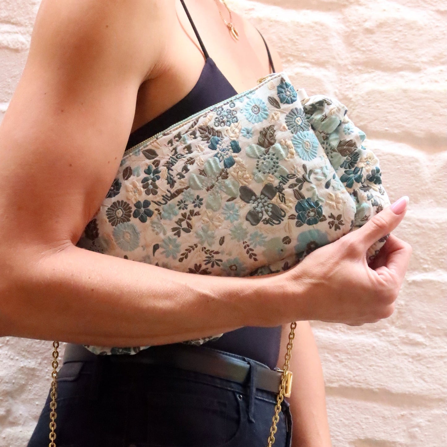 The Dolly Field Flowers Frilly Edge Bag is handcrafted in England from elegant blue and turquoise floral brocade on a cream background, finished with a mint zip and gold hardware. Complete with a white leather wrist strap and bespoke gold chain, it can be worn as a clutch, shoulder or crossbody bag for weddings, festivals, holidays and everyday occasions.