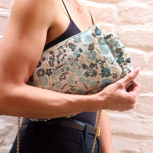 The Dolly Field Flowers Frilly Edge Bag is handcrafted in England from elegant blue and turquoise floral brocade on a cream background, finished with a mint zip and gold hardware. Complete with a white leather wrist strap and bespoke gold chain, it can be worn as a clutch, shoulder or crossbody bag for weddings, festivals, holidays and everyday occasions.