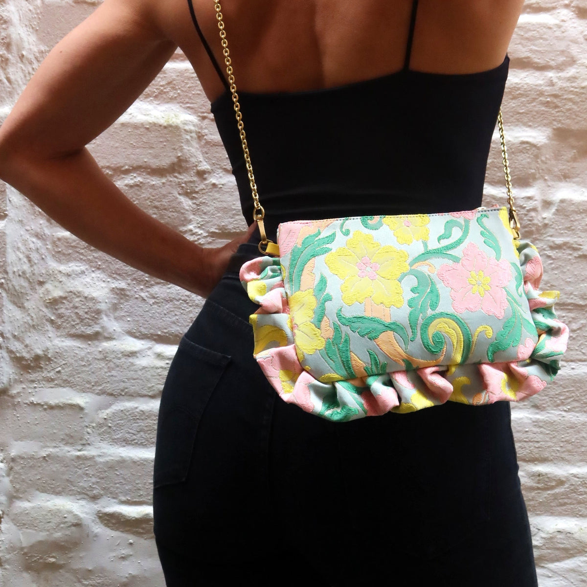 The Dolly Neon Flower Power Frilly Edge Bag is handcrafted in England from vibrant mint green, pink, salmon, yellow and turquoise brocade, finished with a baby blue moiré lining, salmon zip and gold hardware. Complete with a yellow leather wrist strap and bespoke gold chain, it can be worn as a clutch, shoulder or crossbody bag for weddings, festivals, holidays and special occasions.