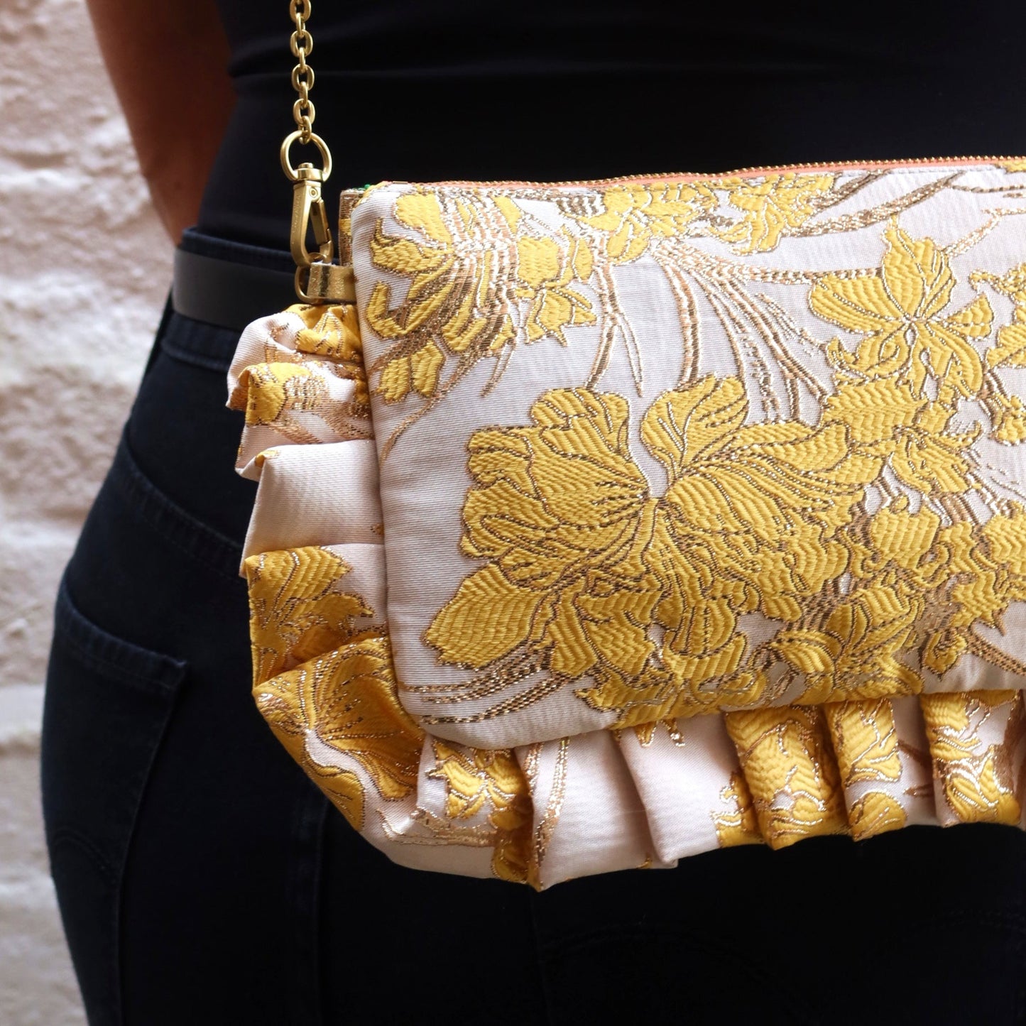 The Dolly Yellow Orchid Frilly Edge Bag is handcrafted in England from luxurious yellow and metallic gold orchid brocade on a cream background, finished with a green moiré-effect silky lining, peach zip and satin gold hardware. Complete with a gold leather wrist strap and bespoke engraved gold chain, it can be worn as a clutch, shoulder or crossbody bag for weddings, festivals, holidays and special occasions.
